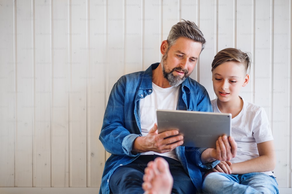 Mature father with small son sitting indoors against white wall, using tablet. Copy space.