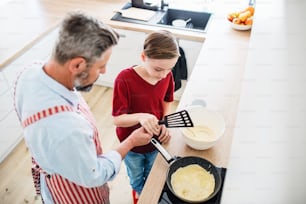 A top view of mature father with small son indoors in kitchen, making pancakes.