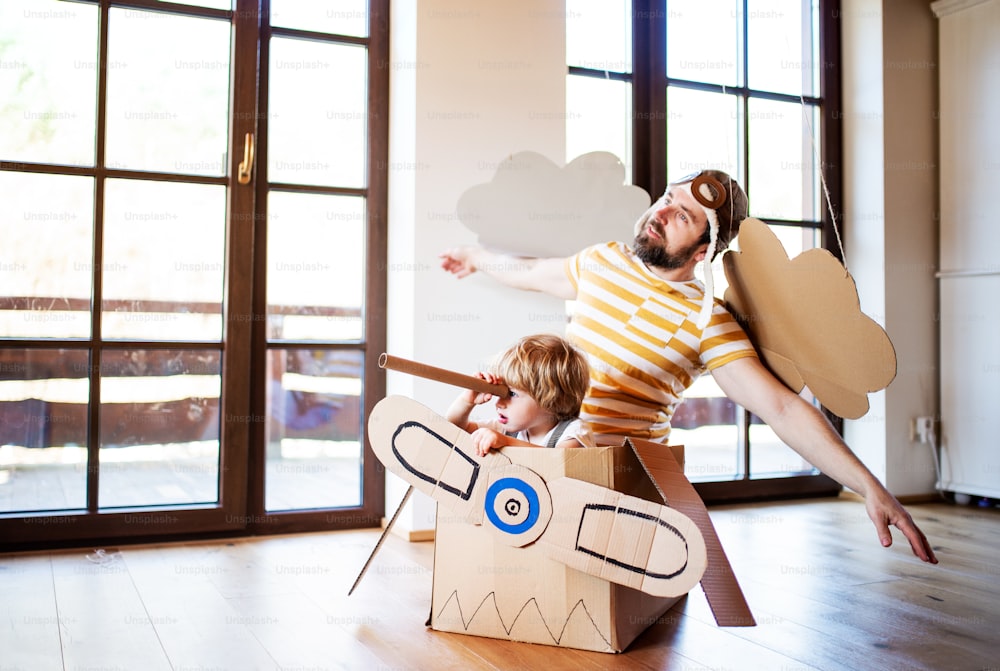 幸せな幼児の少年と父親が、家の屋内で遊んでいるカートン飛行機を持ち、コンセプトを飛ばしている。