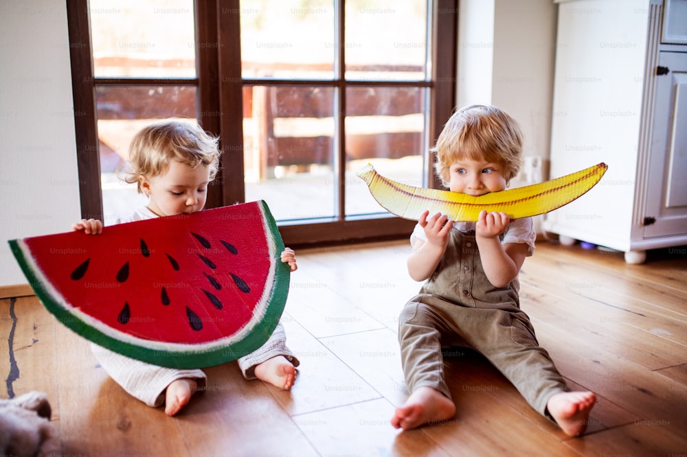 Two toddler children playing with large toy fruit indoors at home, eating fruit concept.