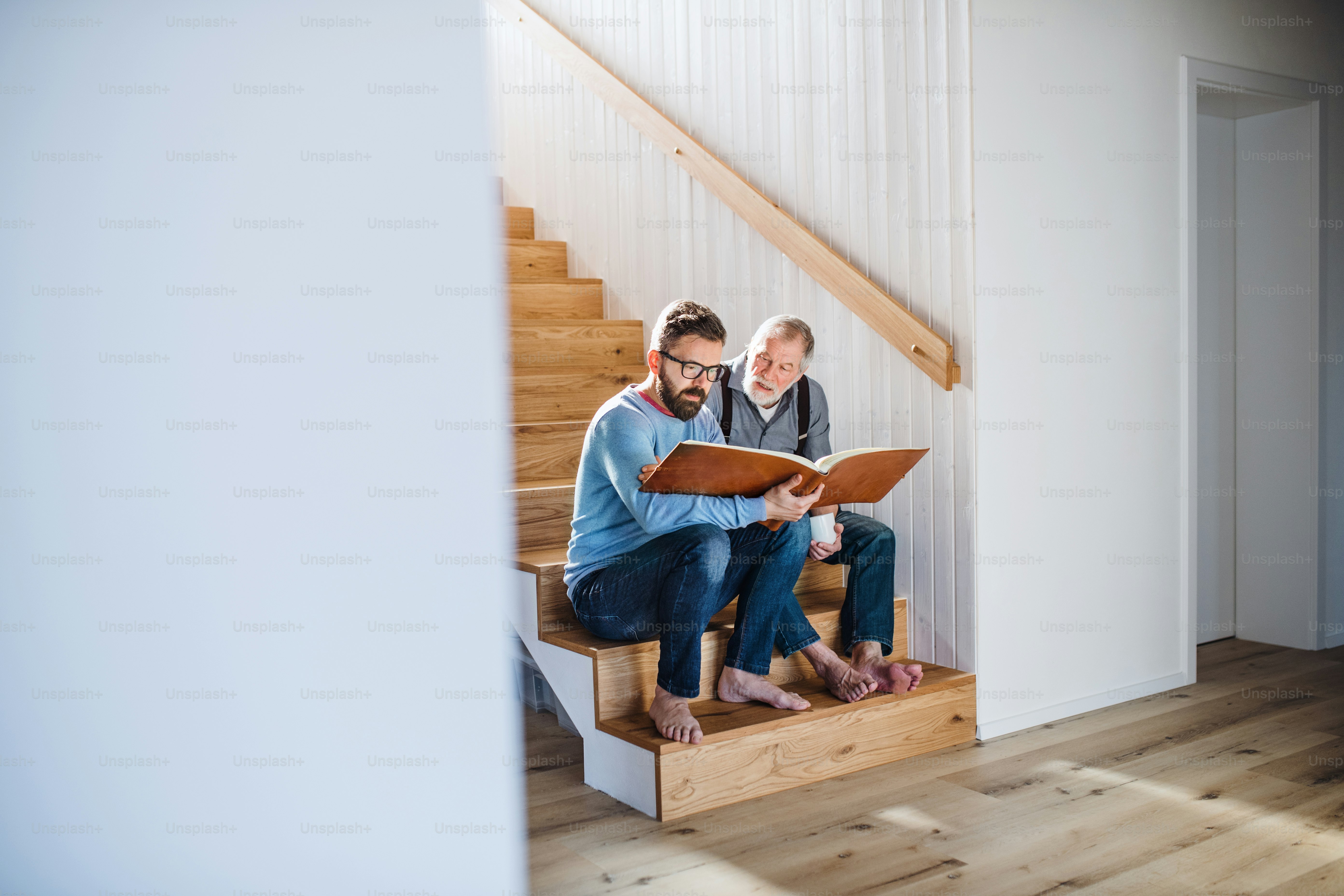 An adult hipster son and senior father sitting on stairs indoors at home, looking at photographs.
