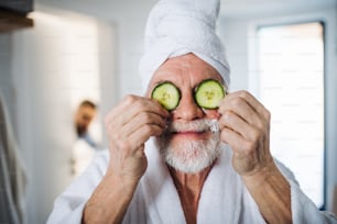 A senior man with cucumber on front of his eyes in bathroom indoors at home.