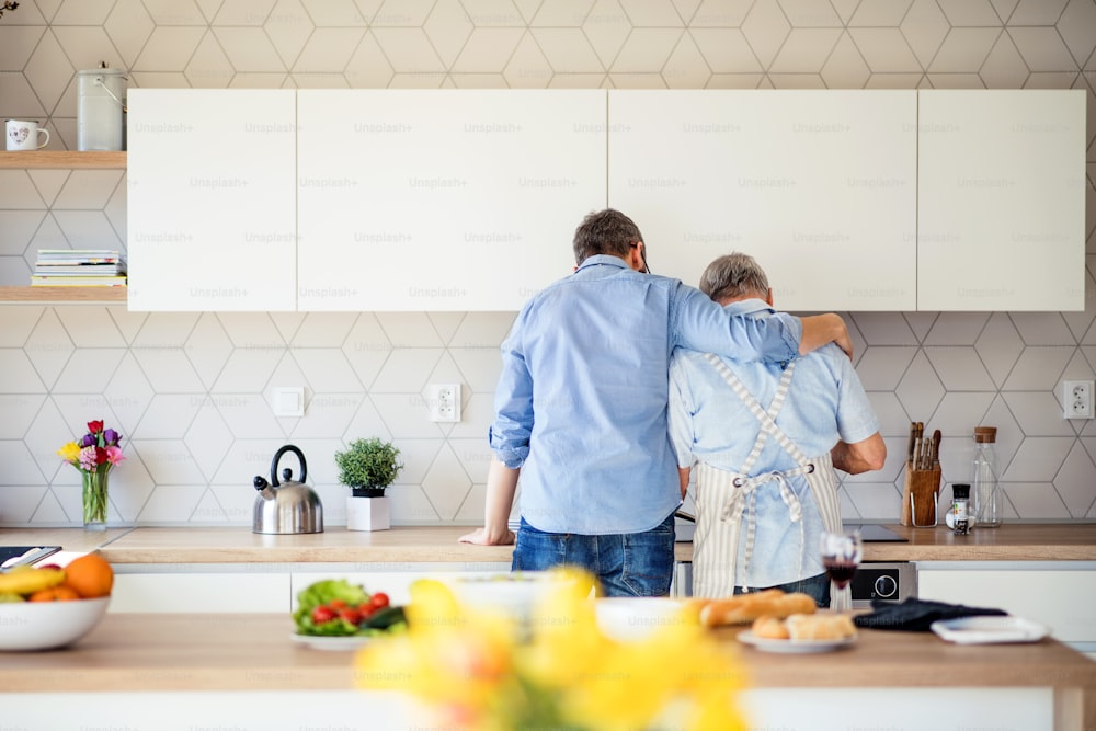 Una vista posteriore del figlio adulto e del padre anziano al chiuso nella cucina di casa, cucinando.