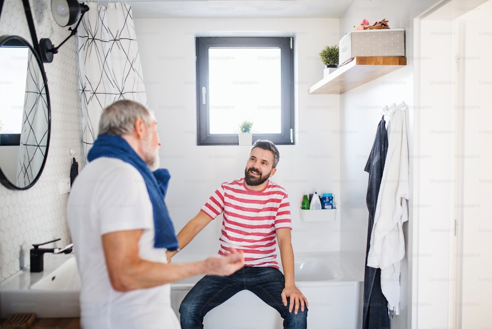 A cheerful adult hipster son and senior father in bathroom indoors at home, talking.
