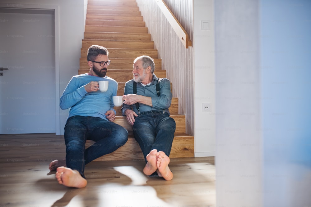 An adult hipster son and senior father with coffee sitting at the bottom of stairs indoors at home, talking.