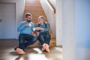 Un hijo hipster adulto y un padre mayor con café sentado en la parte inferior de las escaleras en el interior de casa, hablando.