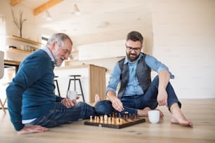 Un fils hipster adulte joyeux et un père aîné assis par terre à l’intérieur à la maison, jouant aux échecs.