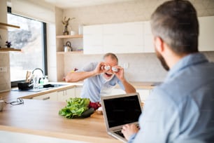 Un fils hipster adulte et un père âgé avec un ordinateur portable à l’intérieur dans la cuisine à la maison, s’amusant.