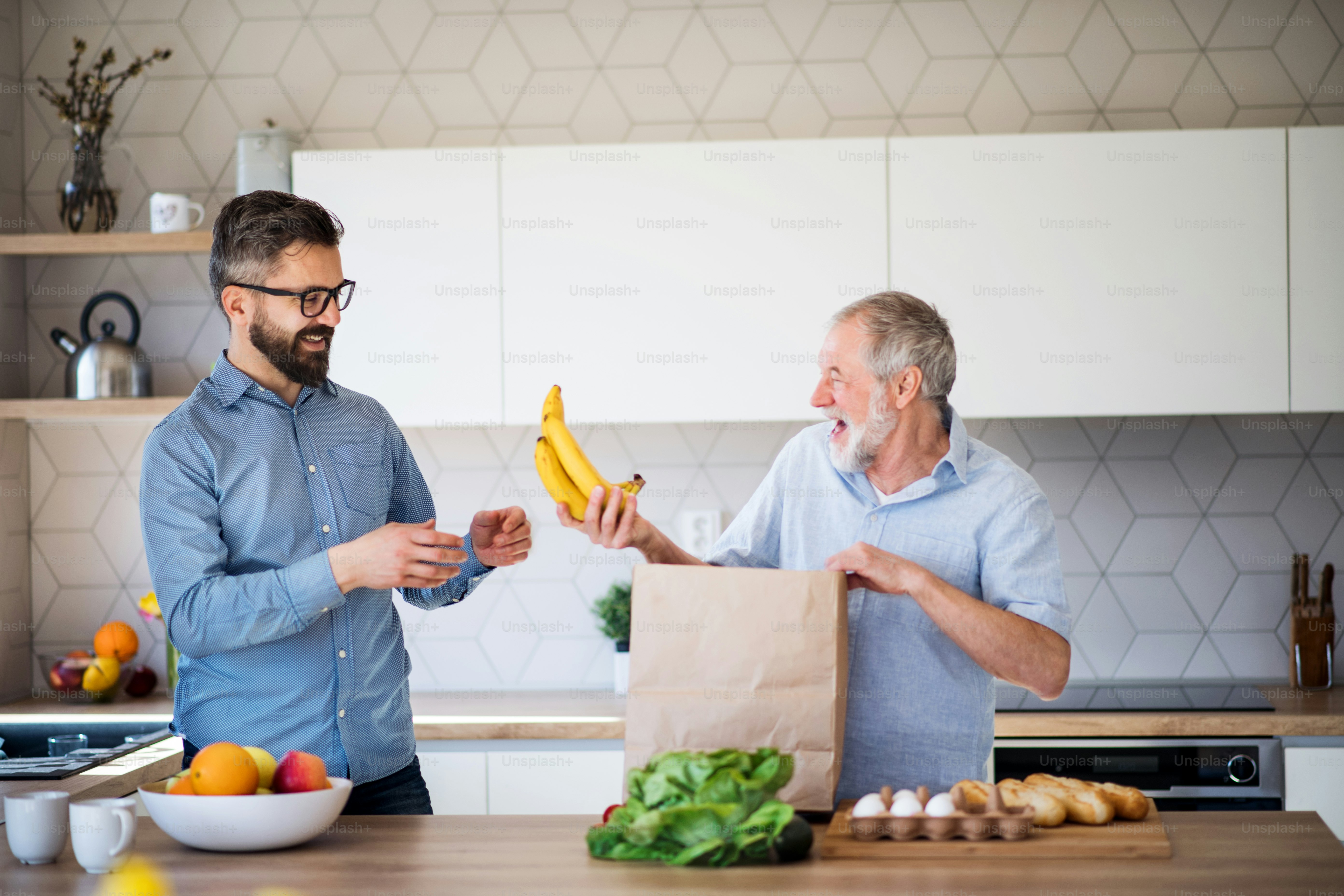 A cheerful adult hipster son and senior father indoors in kitchen at home, unpacking shopping.