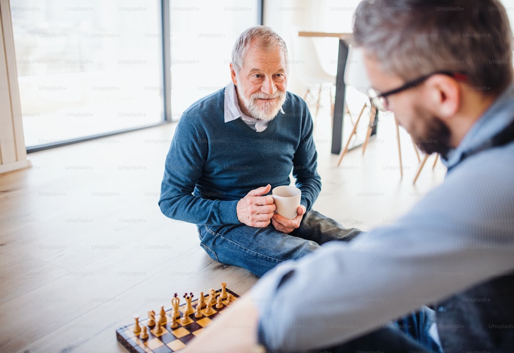Un allegro figlio hipster adulto e padre anziano seduto sul pavimento al chiuso di casa, giocando a scacchi.