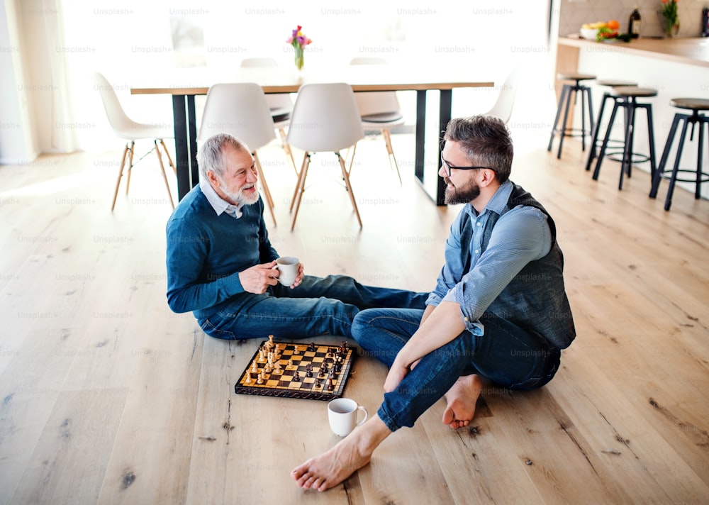 A cheerful adult hipster son and senior father sitting on floor indoors at home, playing chess.