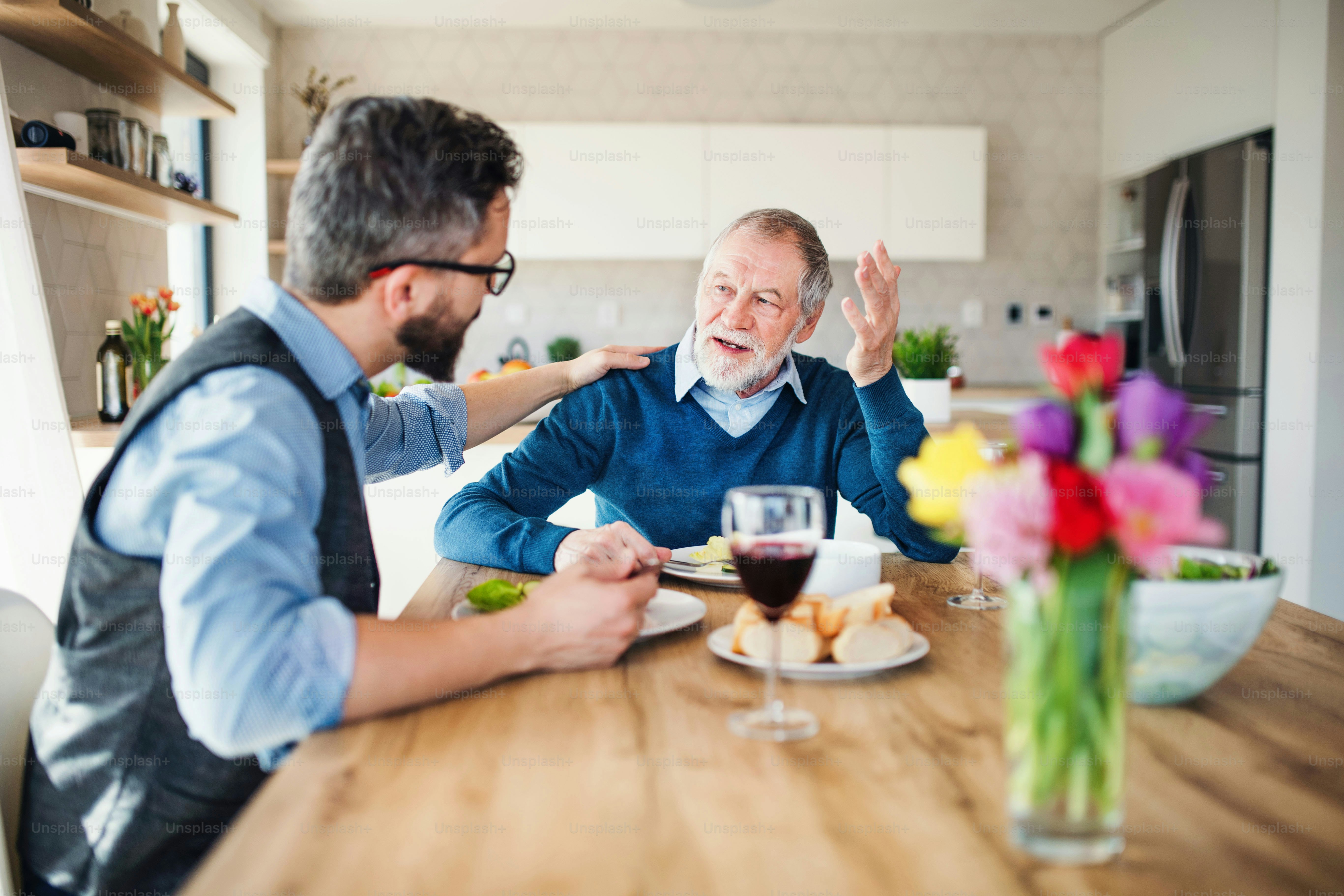 An adult hipster son and senior father sitting at the table indoors at home, eating light lunch.