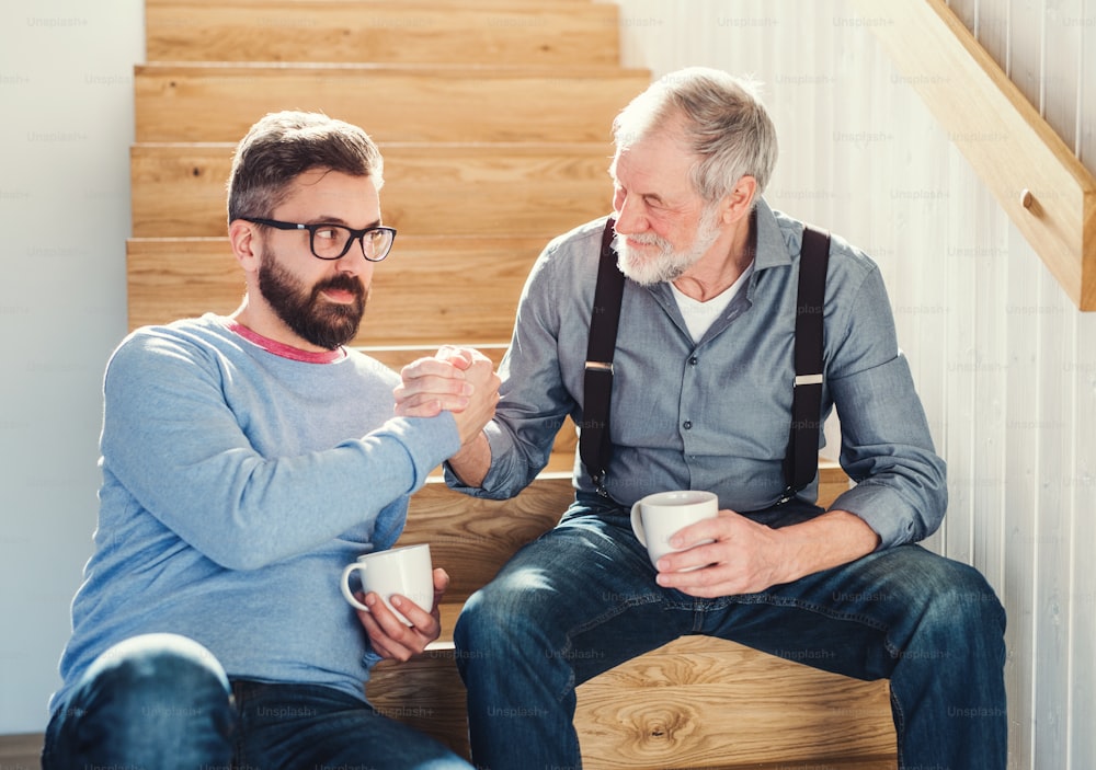 An adult hipster son and senior father with coffee sitting at the bottom of stairs indoors at home, talking.