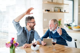 Fils hipster adulte et père âgé avec du vin et une tablette assis à la table à l’intérieur à la maison.