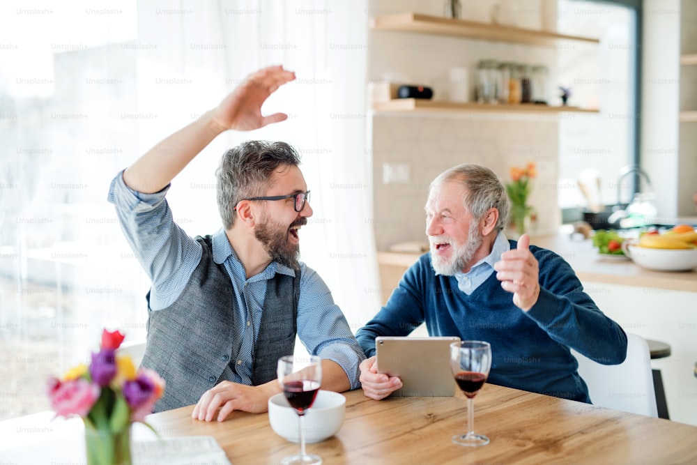 Adult hipster son and senior father with wine and tablet sitting at the table indoors at home.