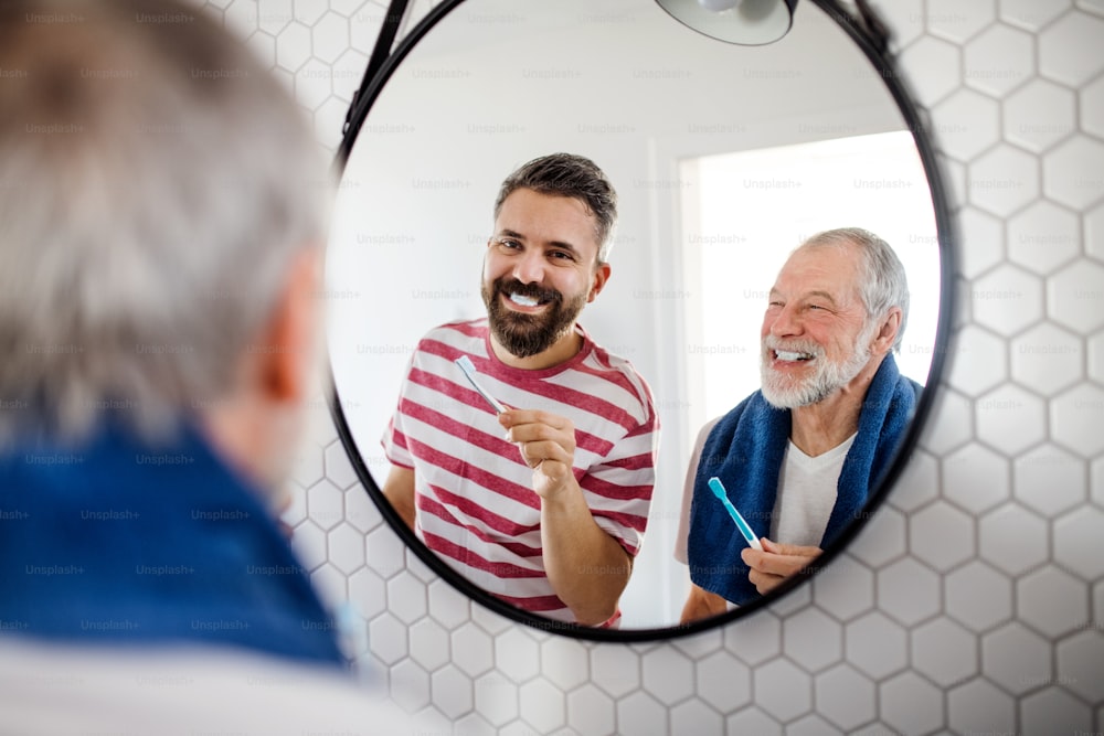 A cheerful adult hipster son and senior father in bathroom indoors at home, looking at mirror and brushing teeth.