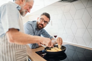 Un fils hipster adulte et un père âgé à l’intérieur dans la cuisine à la maison, en train de cuisiner.