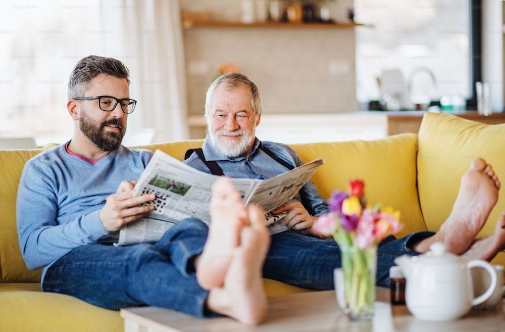 Ein erwachsener Hipster-Sohn und älterer Vater mit Zeitungen, der zu Hause drinnen auf dem Sofa sitzt und sich unterhält.
