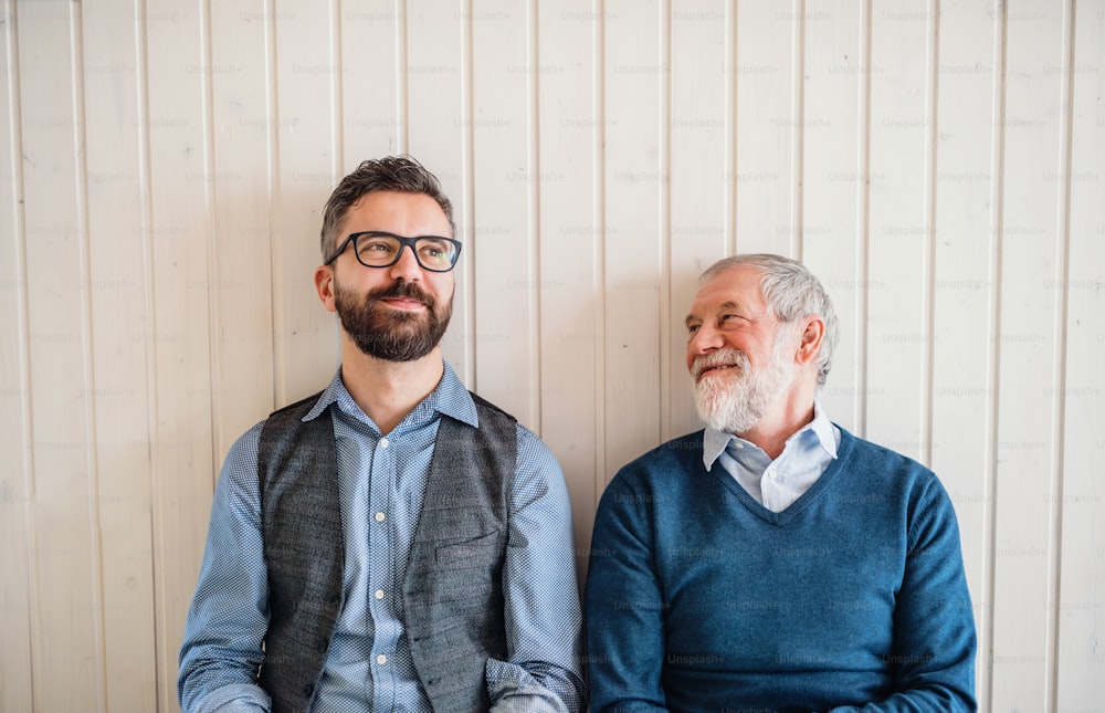 A portrait of adult hipster son and senior father sitting on floor indoors at home, white wooden wall in the background.