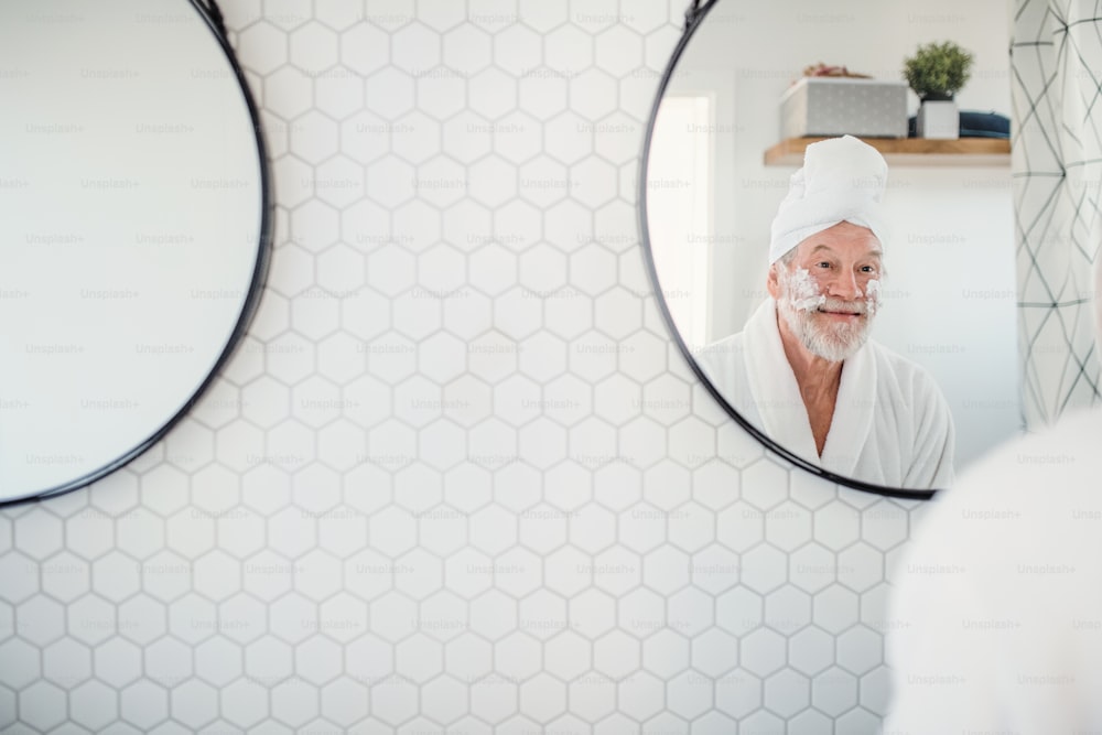 Un hombre mayor haciendo la rutina matutina en el baño en el interior de su casa, mirándose en el espejo. Espacio de copia.