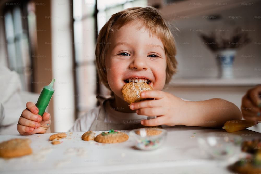 Un petit garçon joyeux assis à table, décorant et mangeant des gâteaux à la maison.