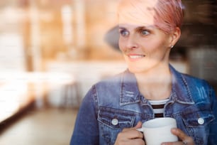 A close-up portrait of young attractive woman with coffee standing by window at home. Shot through glass.