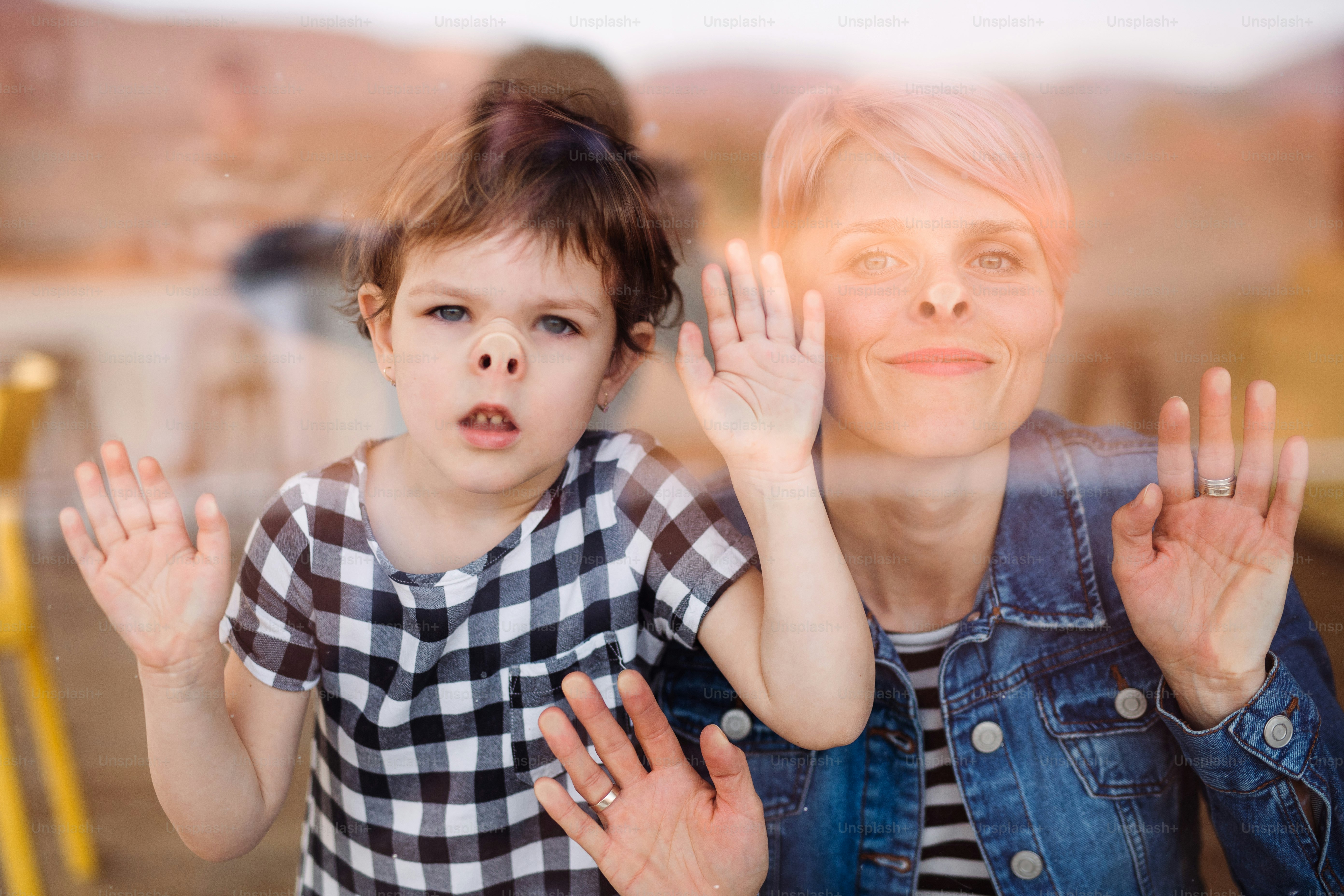 A portrait of young woman with small daughter at home, looking at camera. Shot through glass.