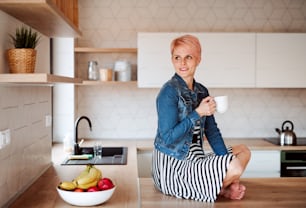 A young attractive woman with a coffee sitting on a counter in a kitchen at home.