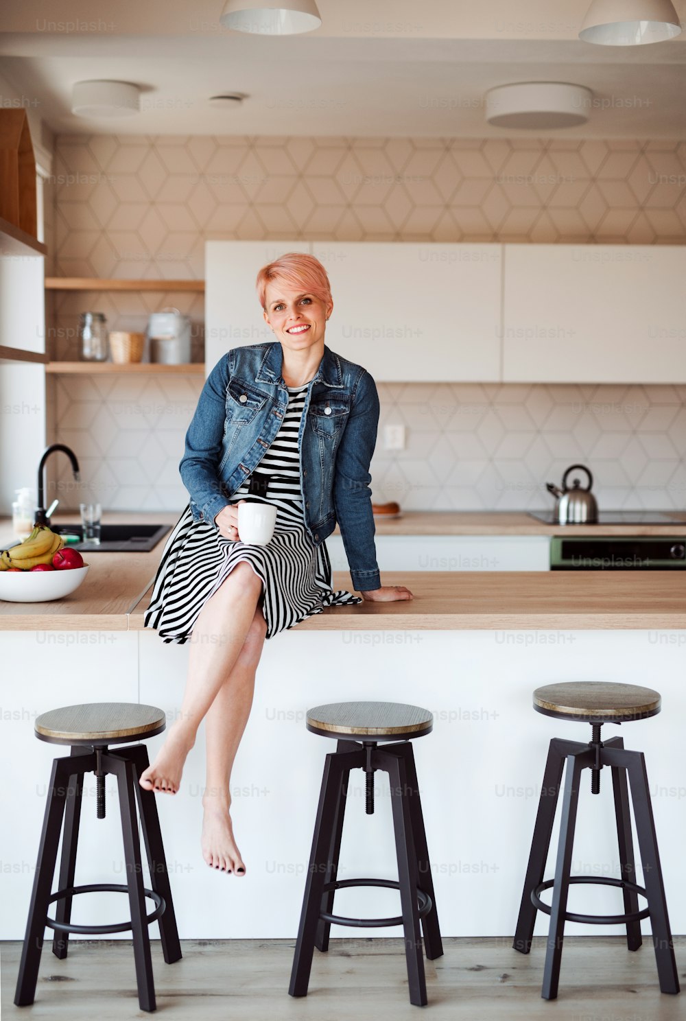 Eine junge attraktive Frau mit einem Kaffee sitzt auf einer Theke in einer Küche zu Hause.