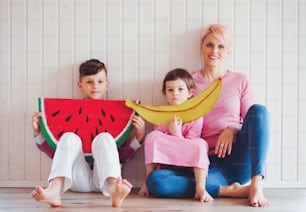 Uma jovem mulher com duas crianças com frutas de brinquedo grandes sentadas no chão em casa.