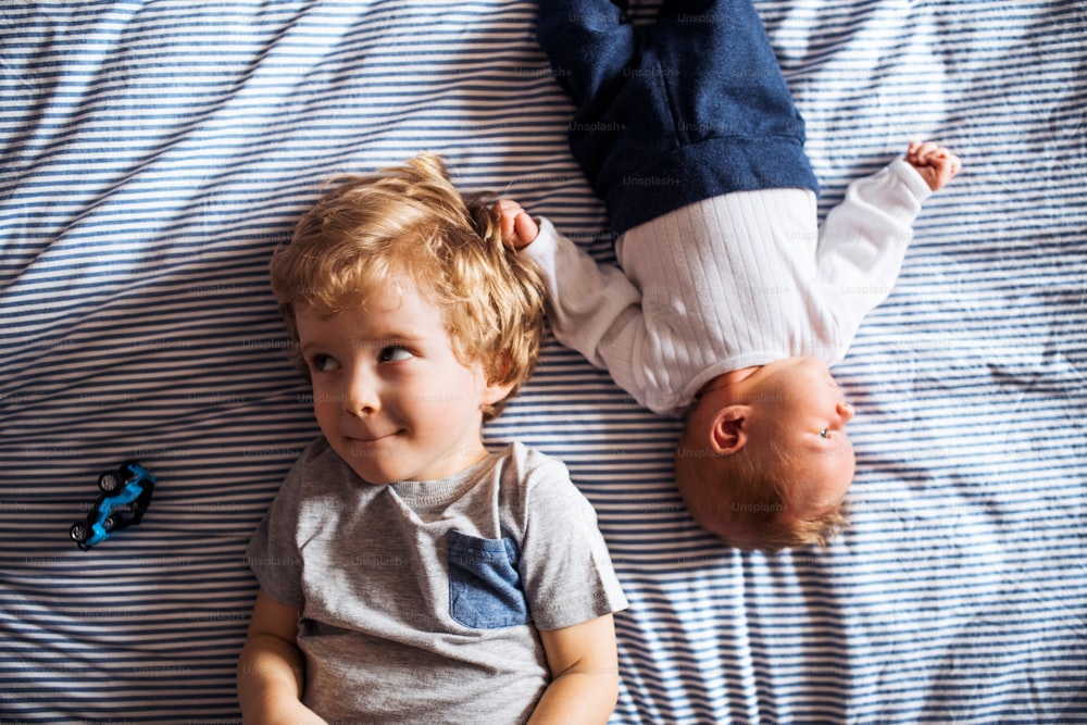 A top view of small toddler boy with a newborn baby brother at home, lying down on bed.