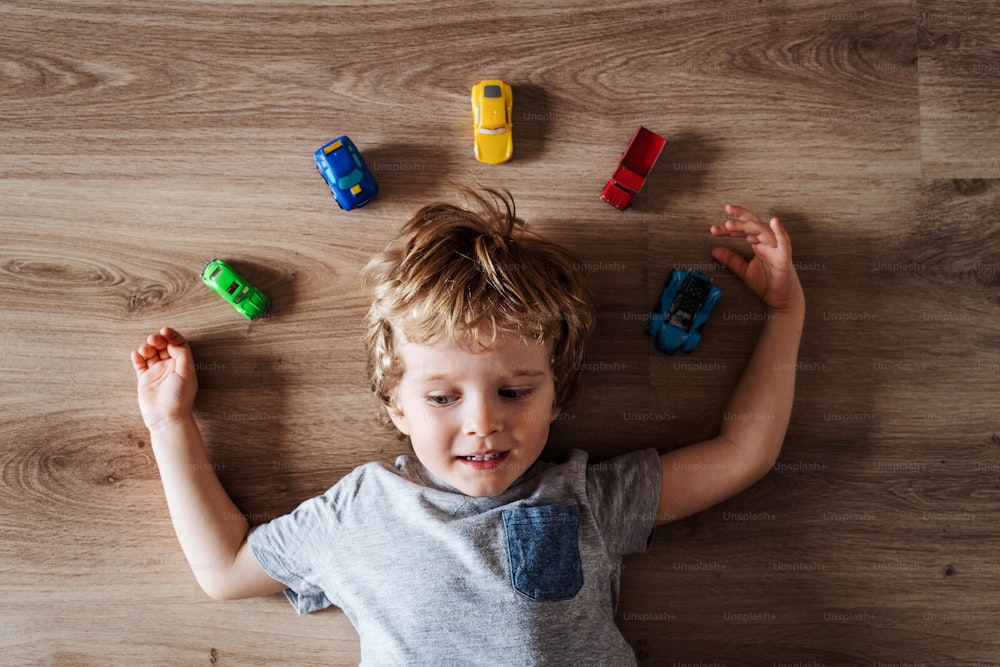 A top view of a toddler boy with toy cars lying on the floor at home.