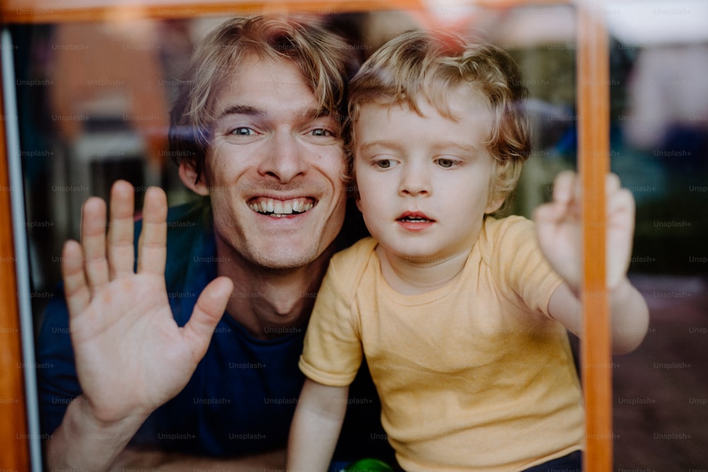 Un padre feliz con un hijo pequeño en casa, mirando a la cámara. Disparado a través del vidrio.