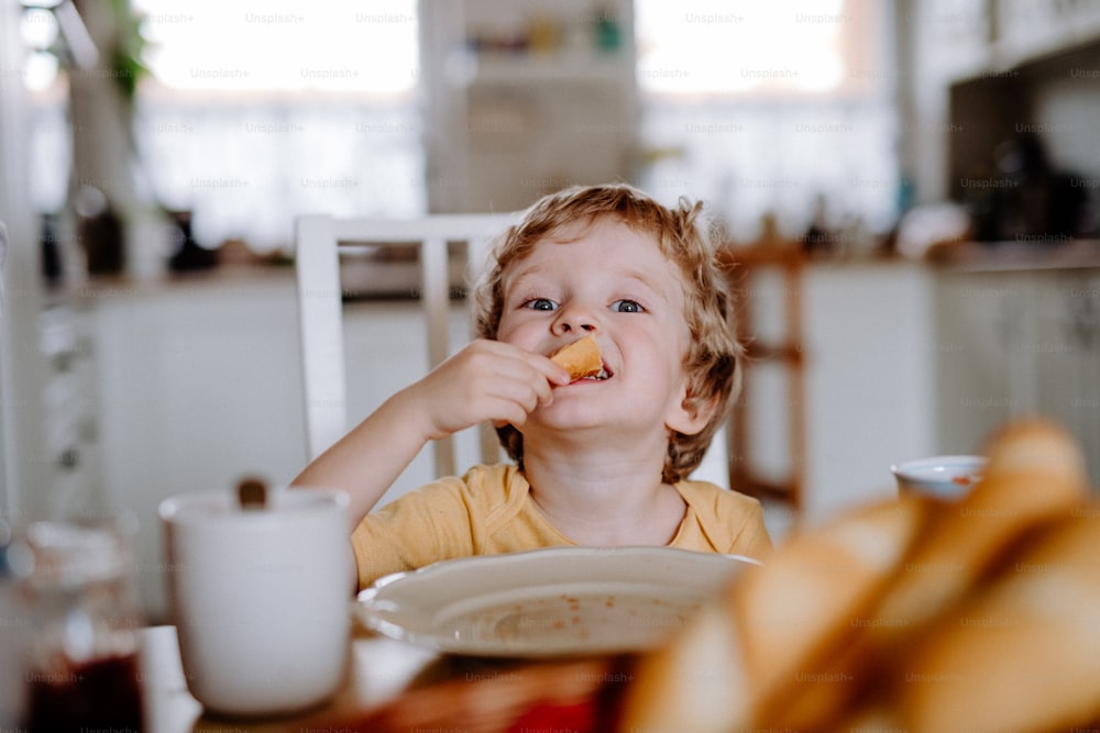 A front view of a happy toddler boy sitting at the table at home, eating.