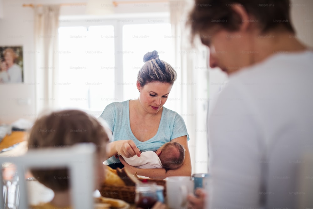 Young parents with a newborn baby and small toddler son sitting at the table at home.