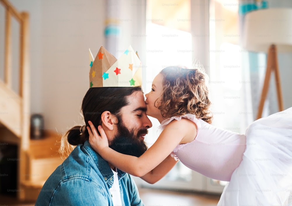 Una vista lateral de una niña pequeña con una corona de princesa y un padre joven en casa, besándose cuando juega.