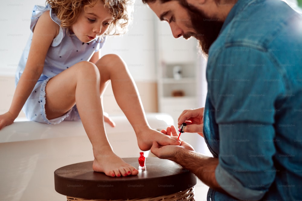 Un jeune père peignant les ongles d’une petite fille dans une salle de bain à la maison.