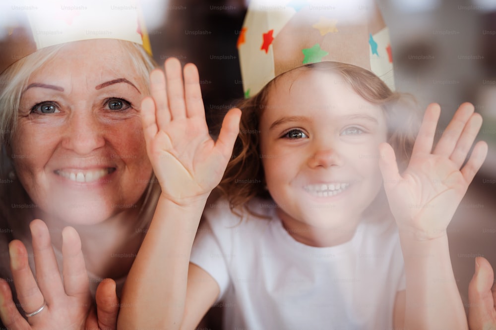 A portrait of small girl and grandmother with paper crown having fun at home. Shot through glass.