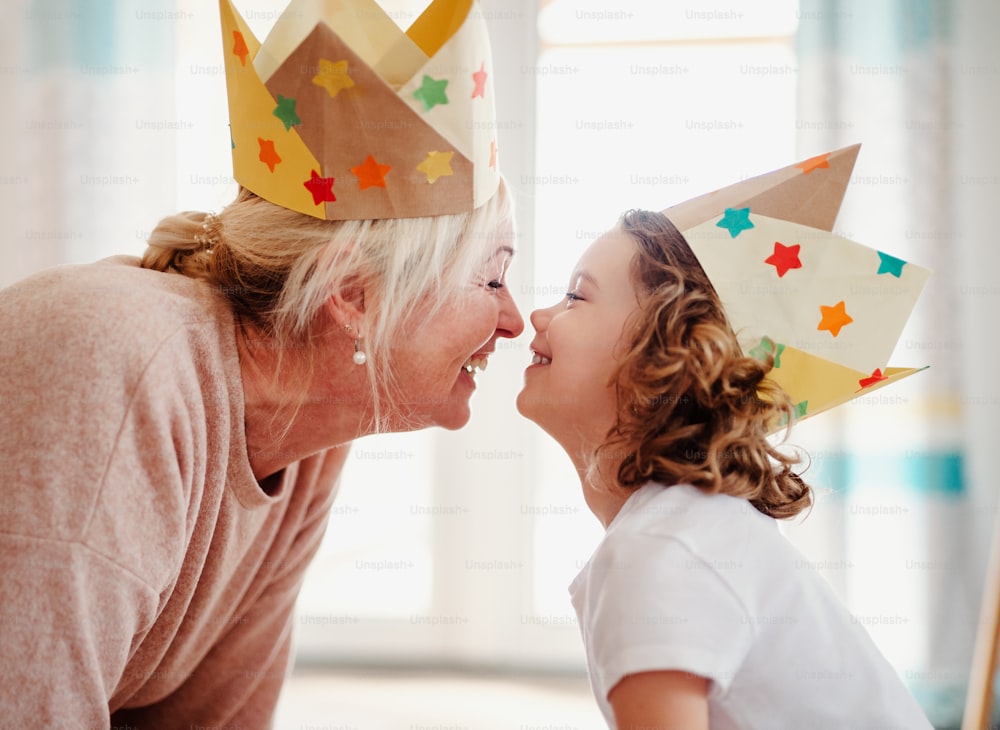 A portrait of small girl and grandmother with paper crown at home, having fun.