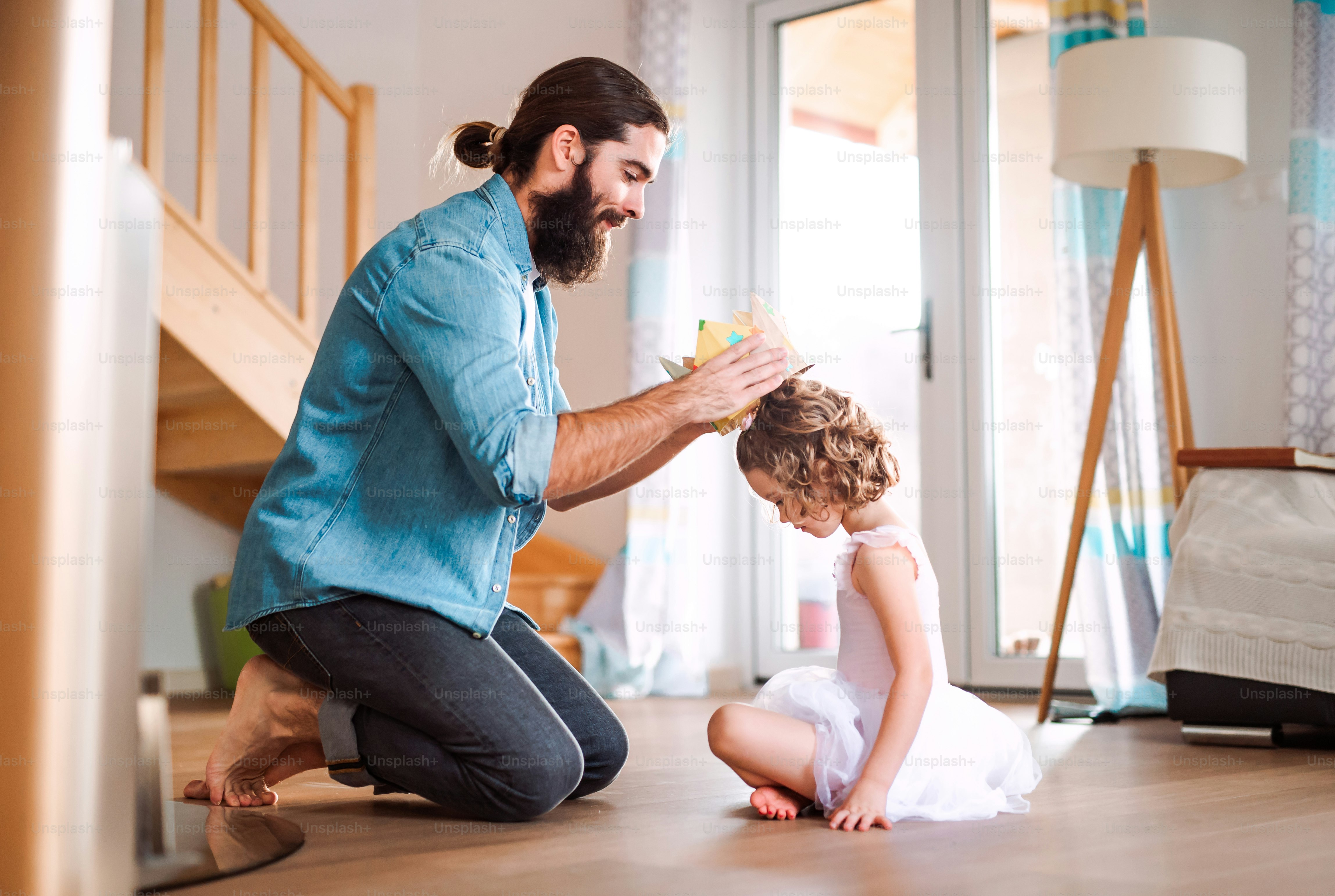 A side view of small girl with a princess crown and young father at home, playing.