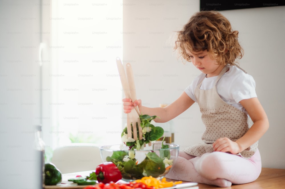 Ein Porträt eines kleinen Mädchens in einer Küche zu Hause, das Gemüsesalat zubereitet.