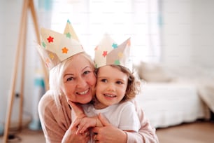 A portrait of small girl and grandmother with paper crown hugging at home.