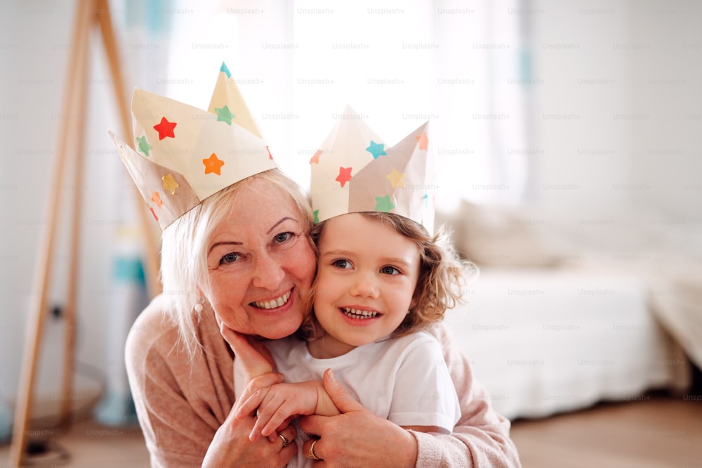 A portrait of small girl and grandmother with paper crown hugging at home.
