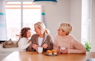 Un retrato de una niña feliz con la madre y la abuela sentadas a la mesa en casa, comiendo magdalenas.