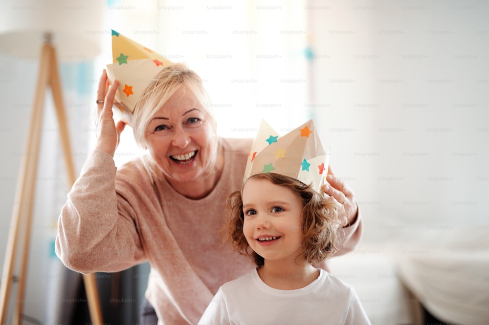 A portrait of small girl and grandmother with paper crown at home, having fun.