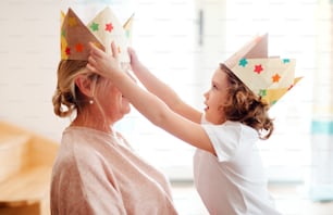 A portrait of small girl and grandmother with paper crown at home, having fun.