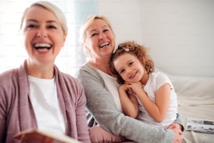 Une petite fille avec mère et grand-mère assise sur un canapé à la maison, en riant.
