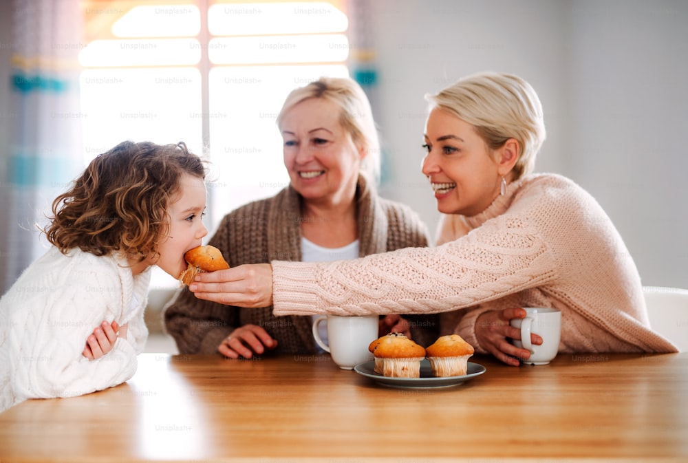어머니와 할머니가 집 식탁에 앉아 머핀을 먹고 있는 행복한 어린 소녀의 초상화.