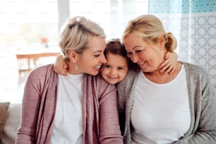 A portrait of happy small girl with mother and grandmother at home, hugging.