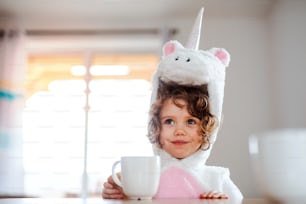 A portrait of happy small girl in unicorn mask sitting at the table at home.